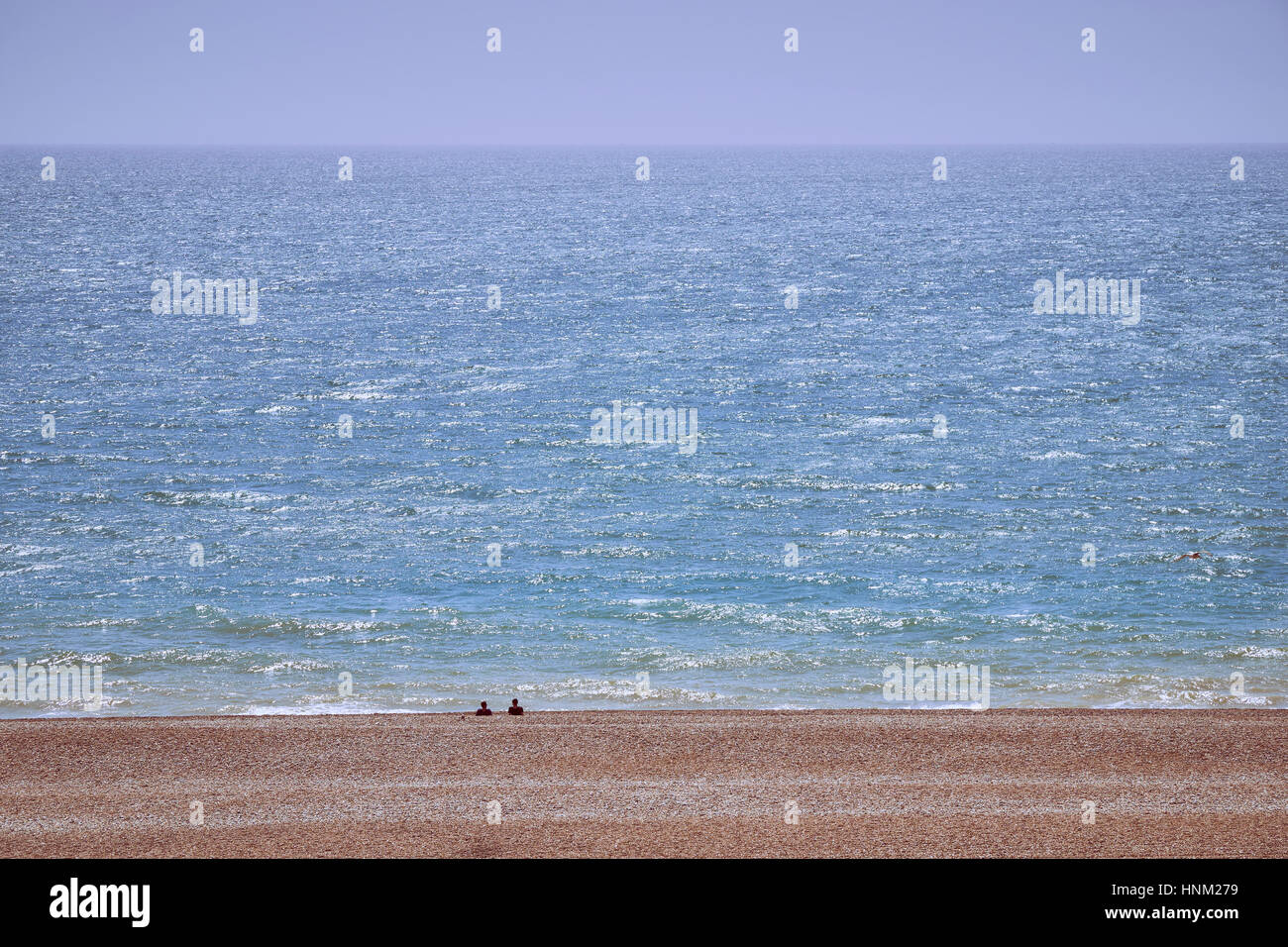 gravel beach and blue sea with two distant people looking out to sea on a sunny summer day Stock Photo