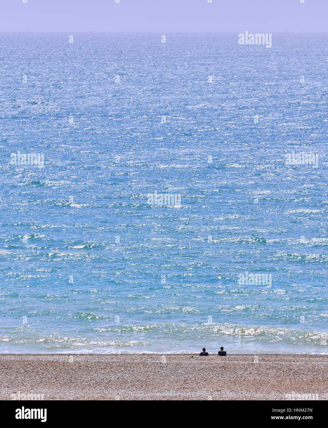 gravel beach and blue sea with two distant people looking out to sea on a sunny summer day Stock Photo