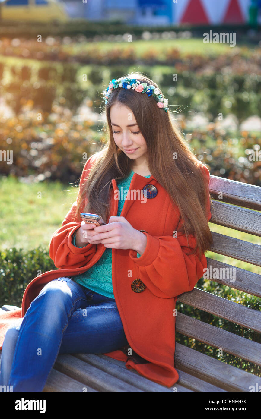 Beautiful young woman in orange coat sitting on the bench with a phone Stock Photo
