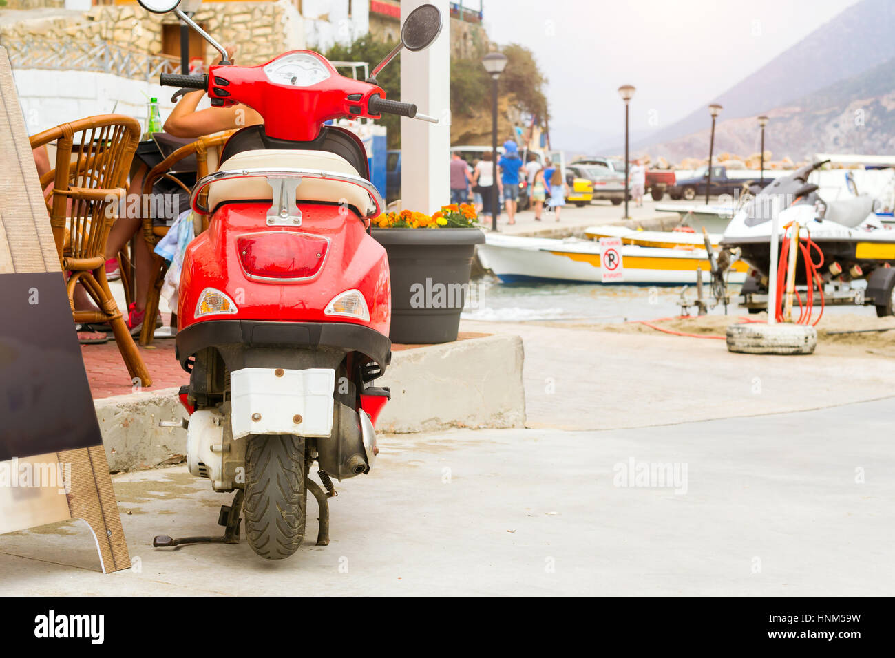 Red two-wheeled moped parked on Mithos beach promenade in sea bay of resort village Bali. Classic Greek stone architecture of resort, tourists walk st Stock Photo