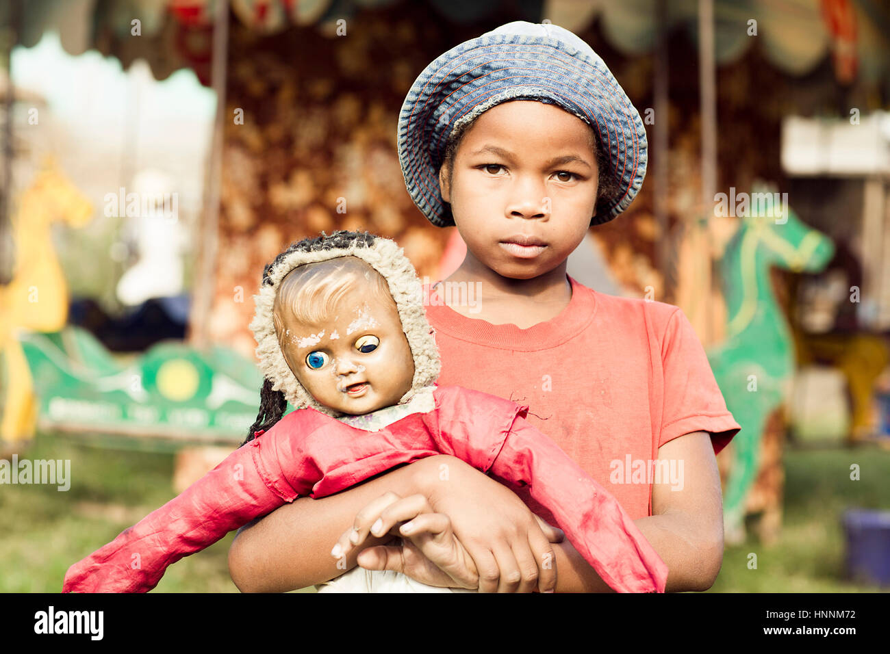Young malagasy girl pose with her worn-out doll in the front of the carousel in the amusement park in Madagascar Stock Photo