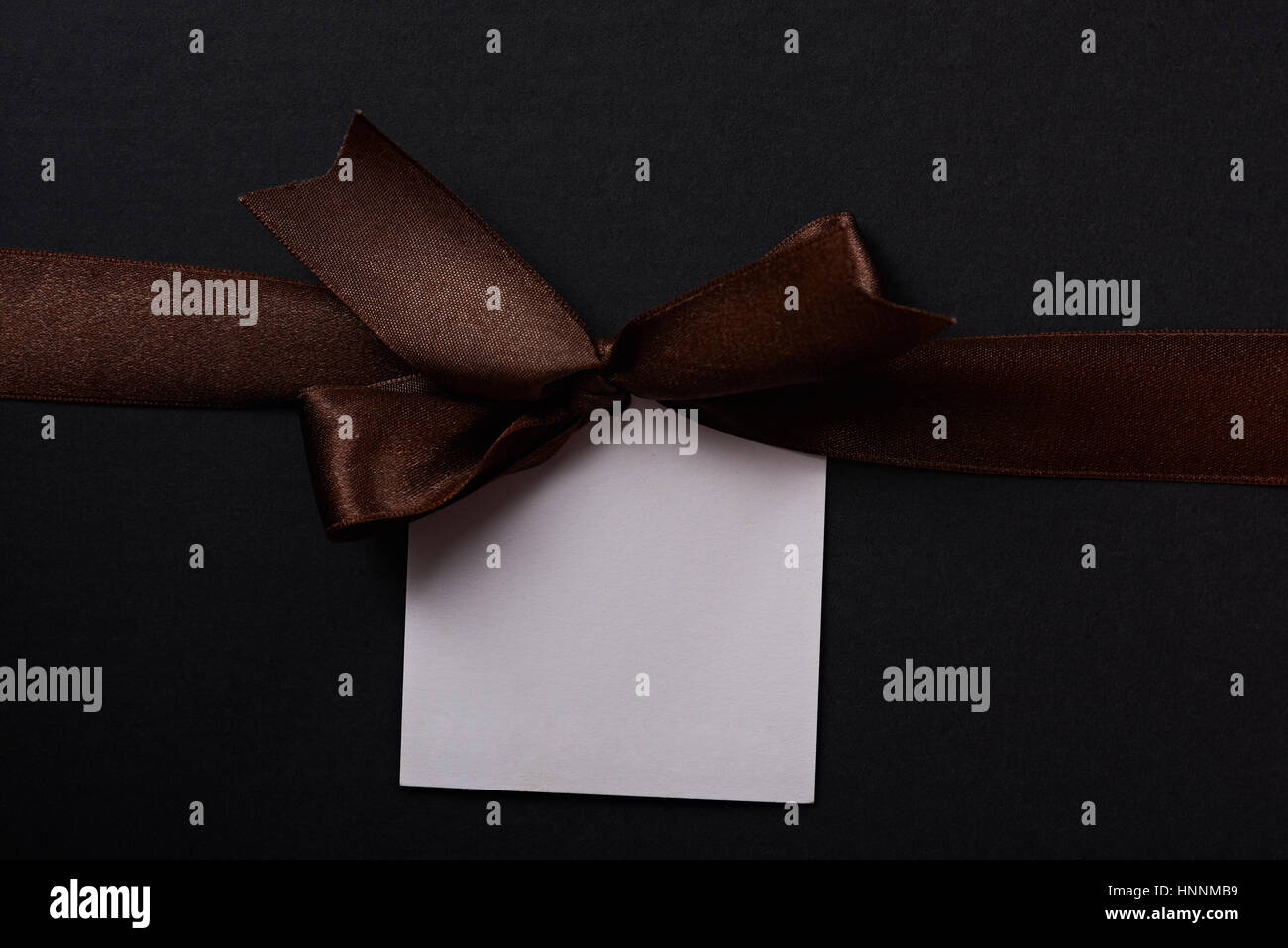 Brown gift bow with paper white label tag on dark background. Brown ribbon with empty tag Stock Photo