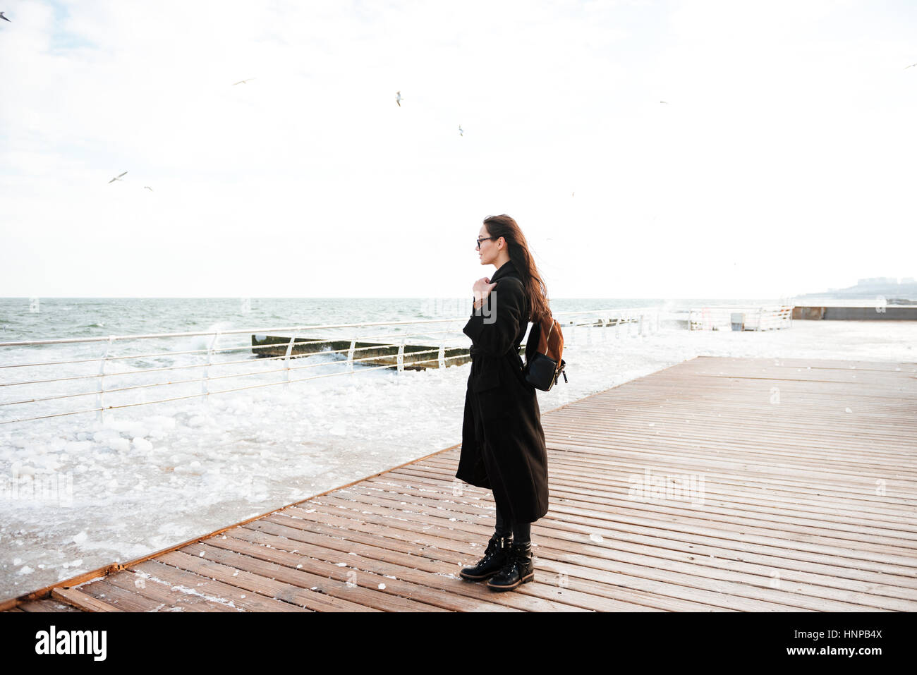Beautiful young woman standing on pier and looking at the sea in winter Stock Photo