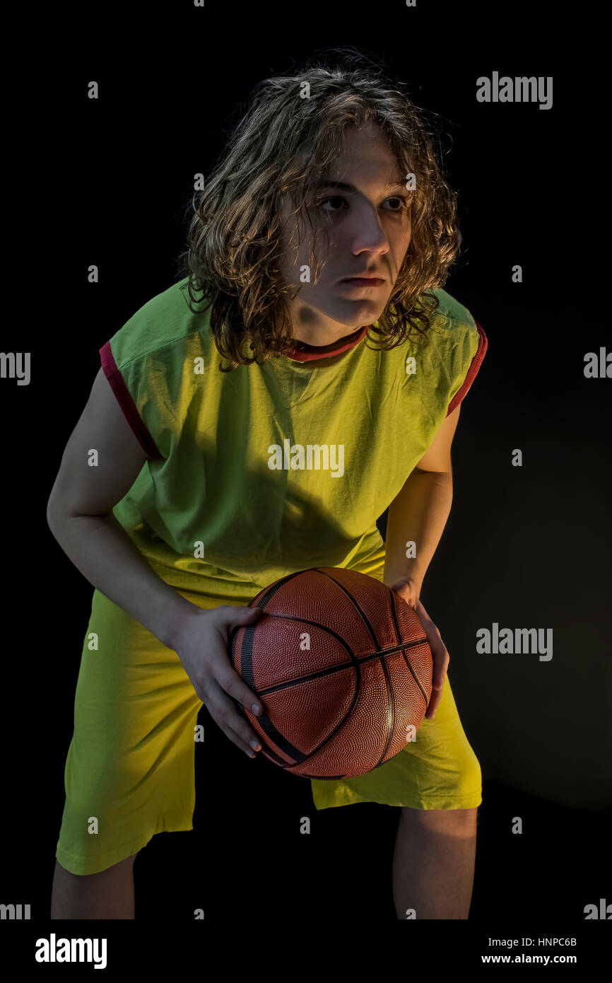 Young boy with long blond hair wearing a green jersey in a squatting position and holding a basketball focusing on the making the shot. Stock Photo