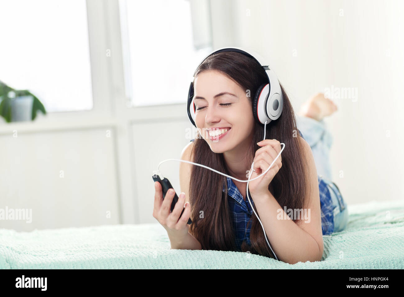 Happy relaxed girl listening music using headphones and smartphone lying on bed at home. People, leisure and technology concept - happy woman with clo Stock Photo