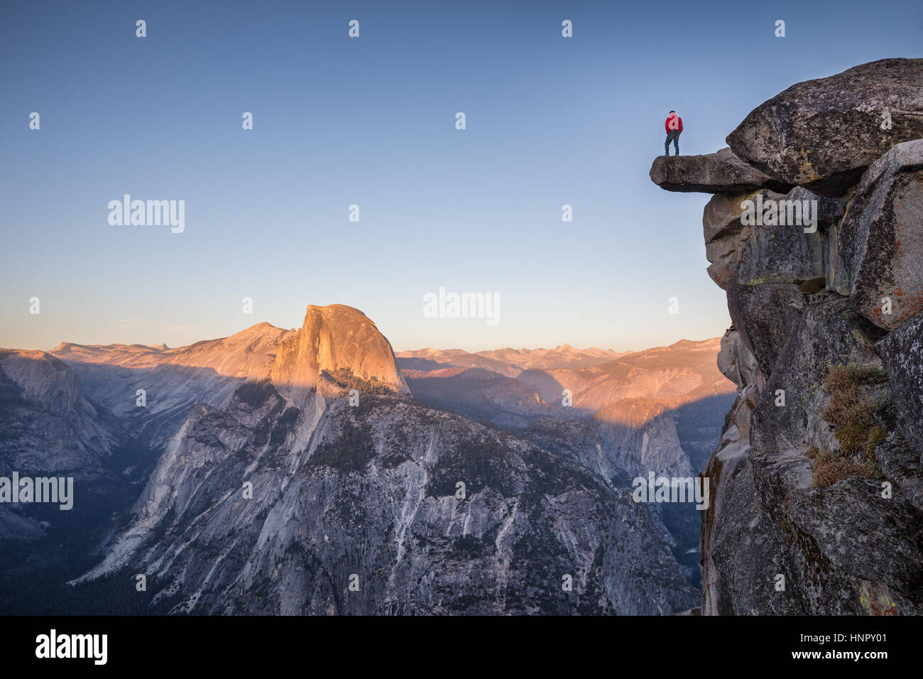 A male hiker standing on an overhanging rock at Glacier Point enjoying the breathtaking view towards famous Half Dome in beautiful twilight at sunset Stock Photo