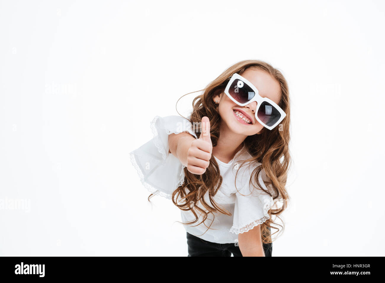 Cheerful little girl in sunglasses smiling and showing thumbs up over white background Stock