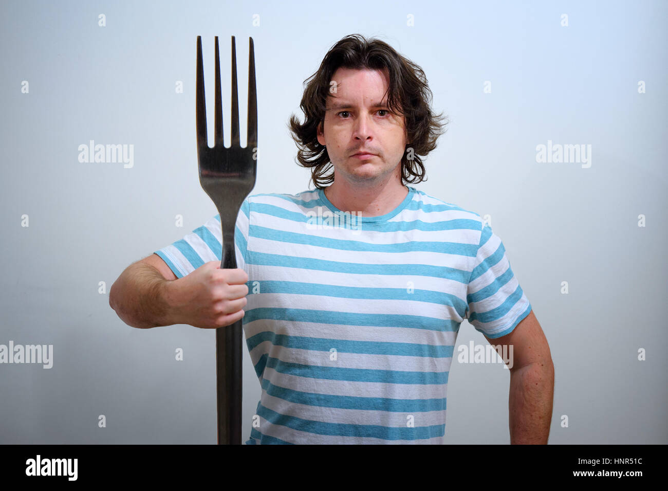 White man in blue and white striped t-shirt with big fork in hand as a Neptune trident. Conceptual foto with fantasy aspect for food, healthy style an Stock Photo