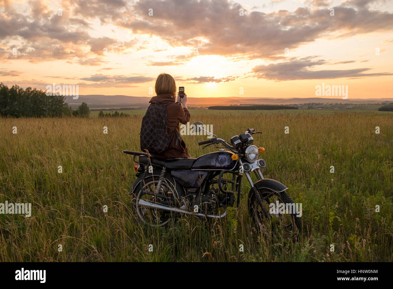 Caucasian woman with motorcycle in field photographing sunset Stock Photo