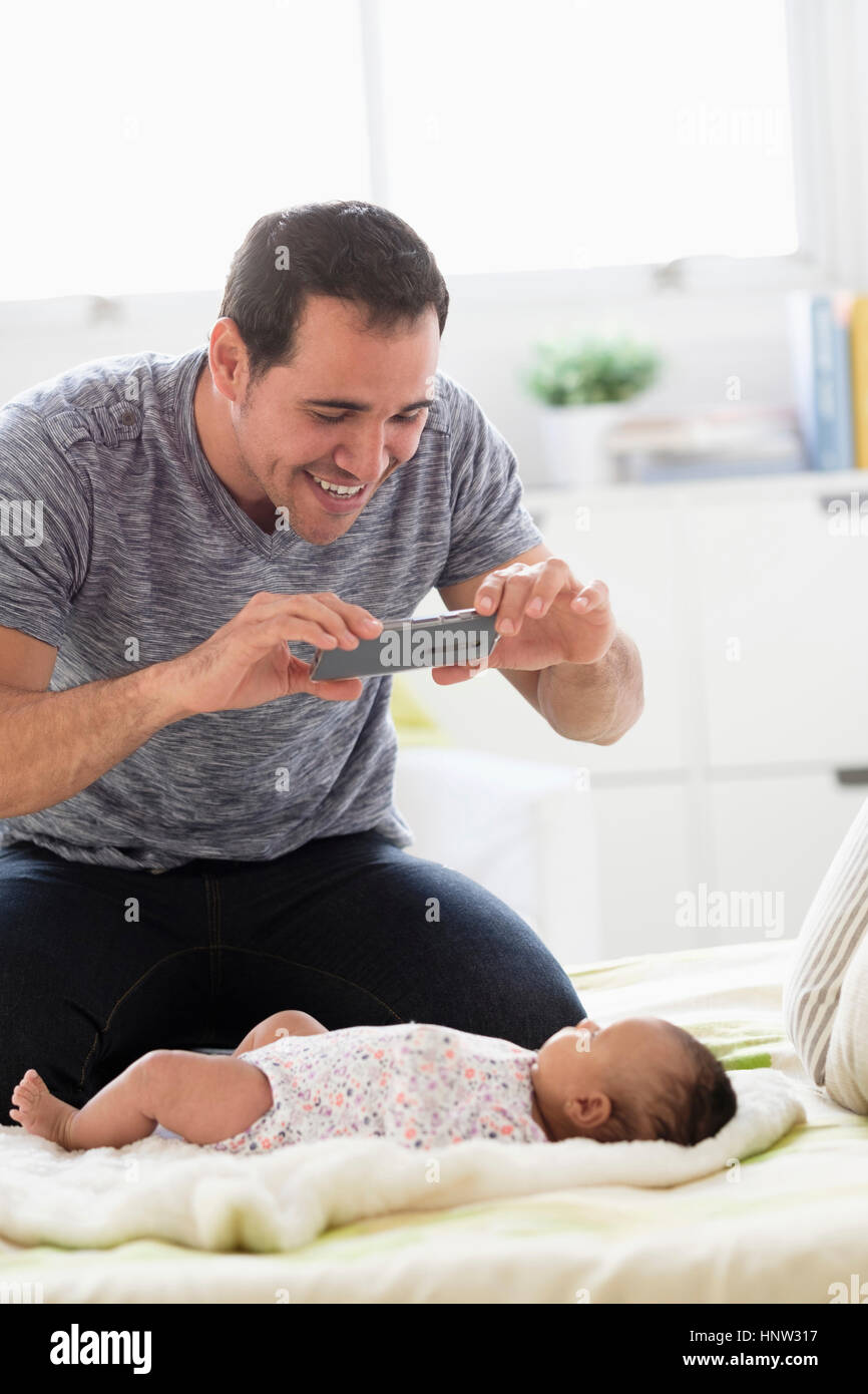 Hispanic father on bed photographing baby daughter with cell phone Stock Photo
