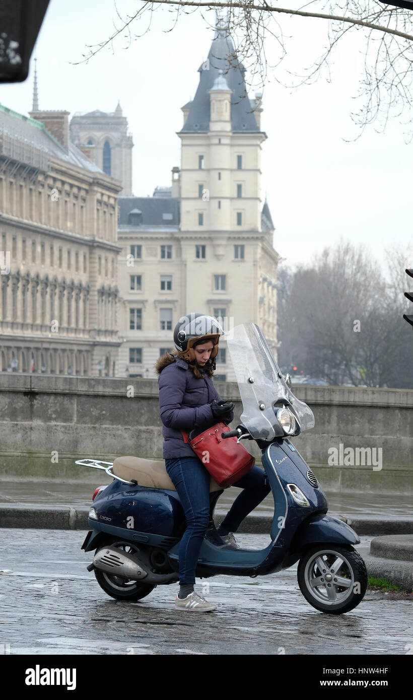 A young woman checks her phone as she waits at a set of traffic lights on her Vespa scooter at Post Neuf, Paris. Stock Photo