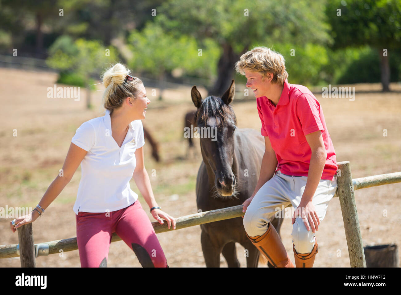 Young male and female grooms sitting on paddock fence at rural stables Stock Photo