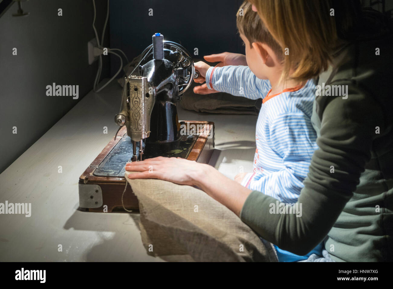 Over shoulder view of boy with mother learning to turn sewing machine ...