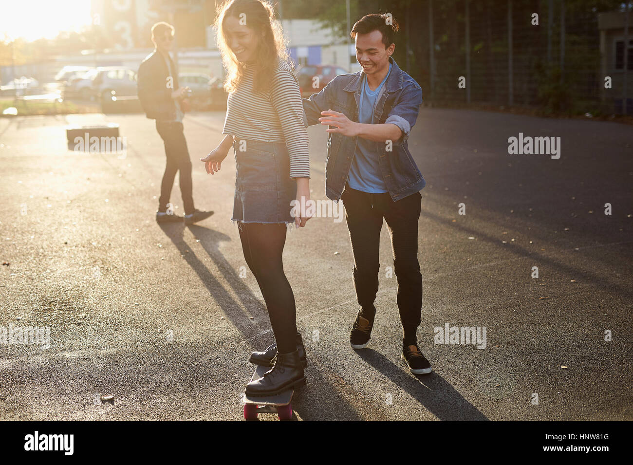 Young man pushing young female skateboarder on sunlit street Stock Photo