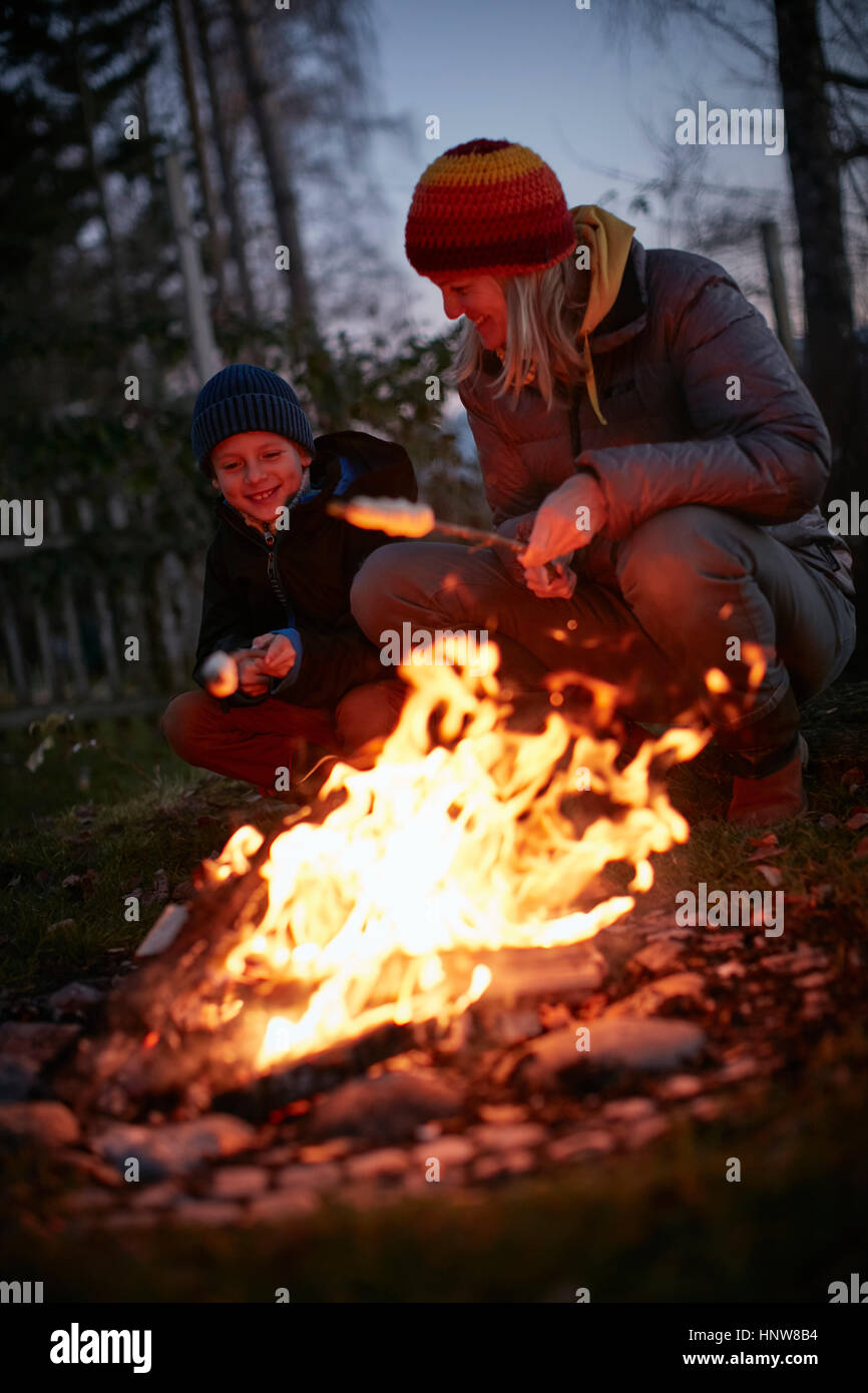 Mature woman and son toasting marshmallows on garden campfire at dusk Stock Photo
