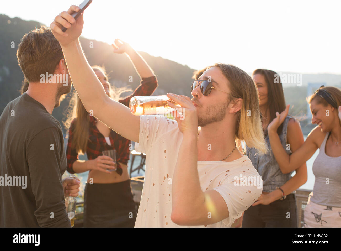Young man taking selfie drinking beer at waterfront roof terrace party, Budapest, Hungary Stock Photo