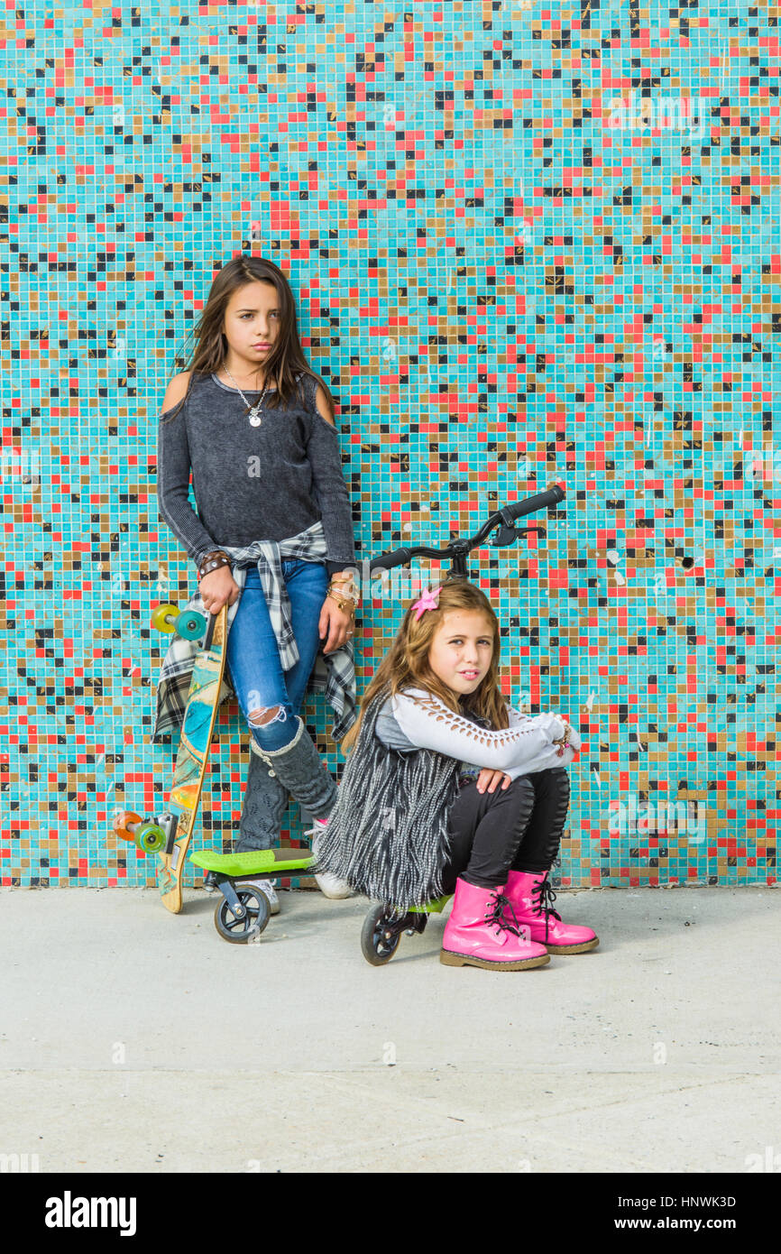 Portrait of two sisters with skateboards and scooter by multi-coloured urban wall Stock Photo