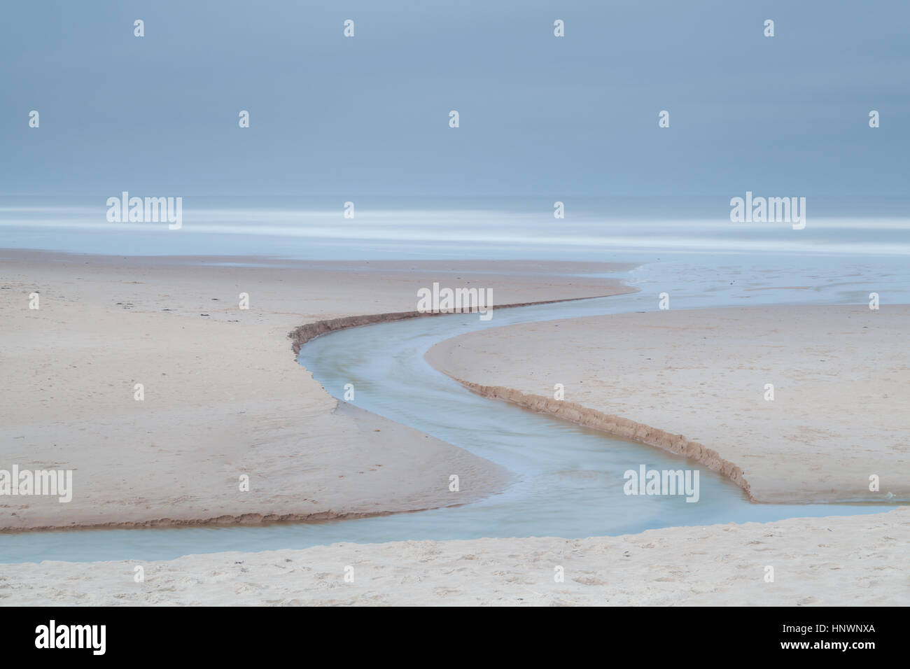 S curve of a stream as it flows across the golden sands of Druridge Bay into the North Sea, Northumberland in the blue hour Stock Photo