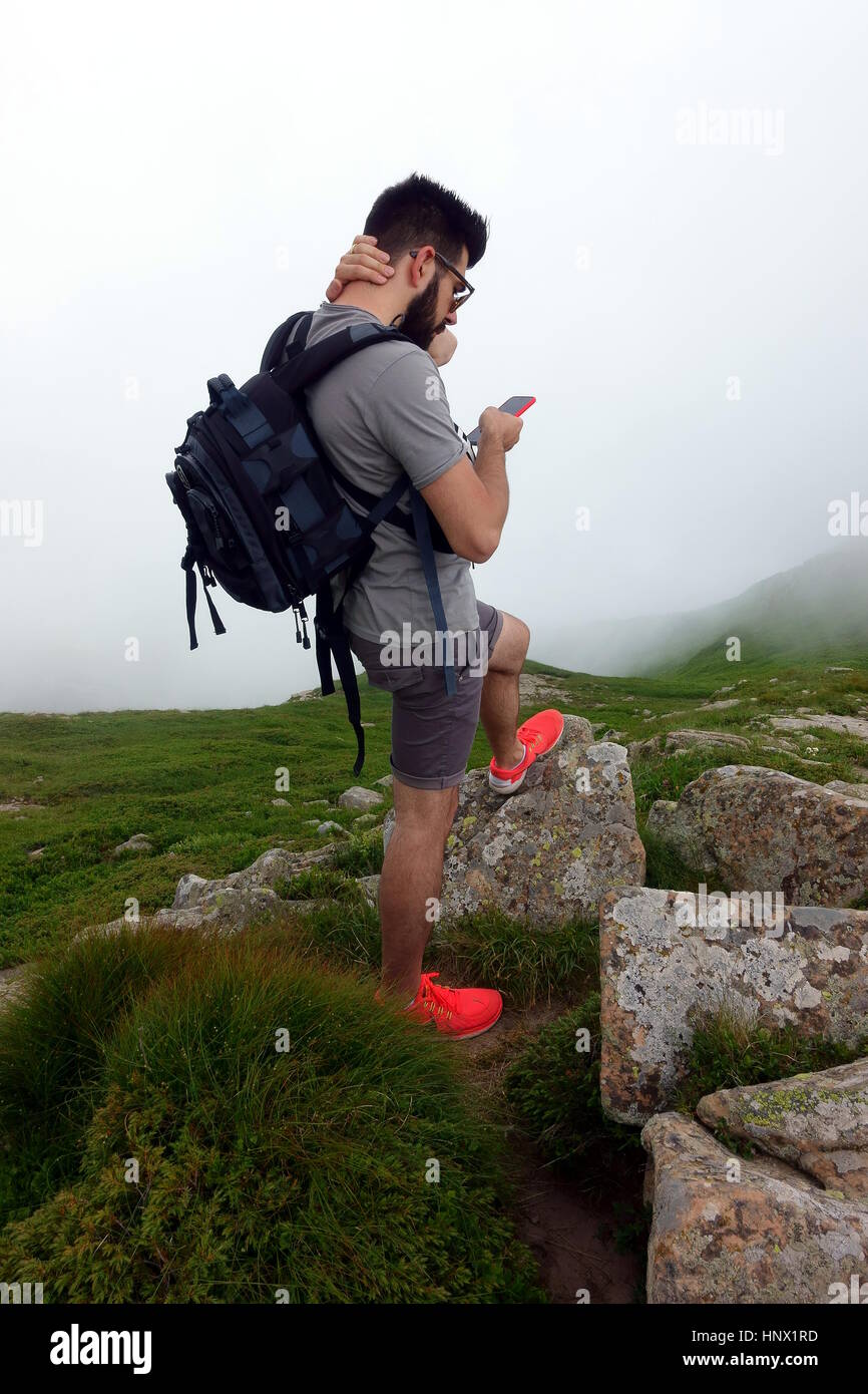 Lost in the clouds - Looking for direction in the fog - Young man with daypack against a misty mountain landscape finding direction with mobile phone Stock Photo