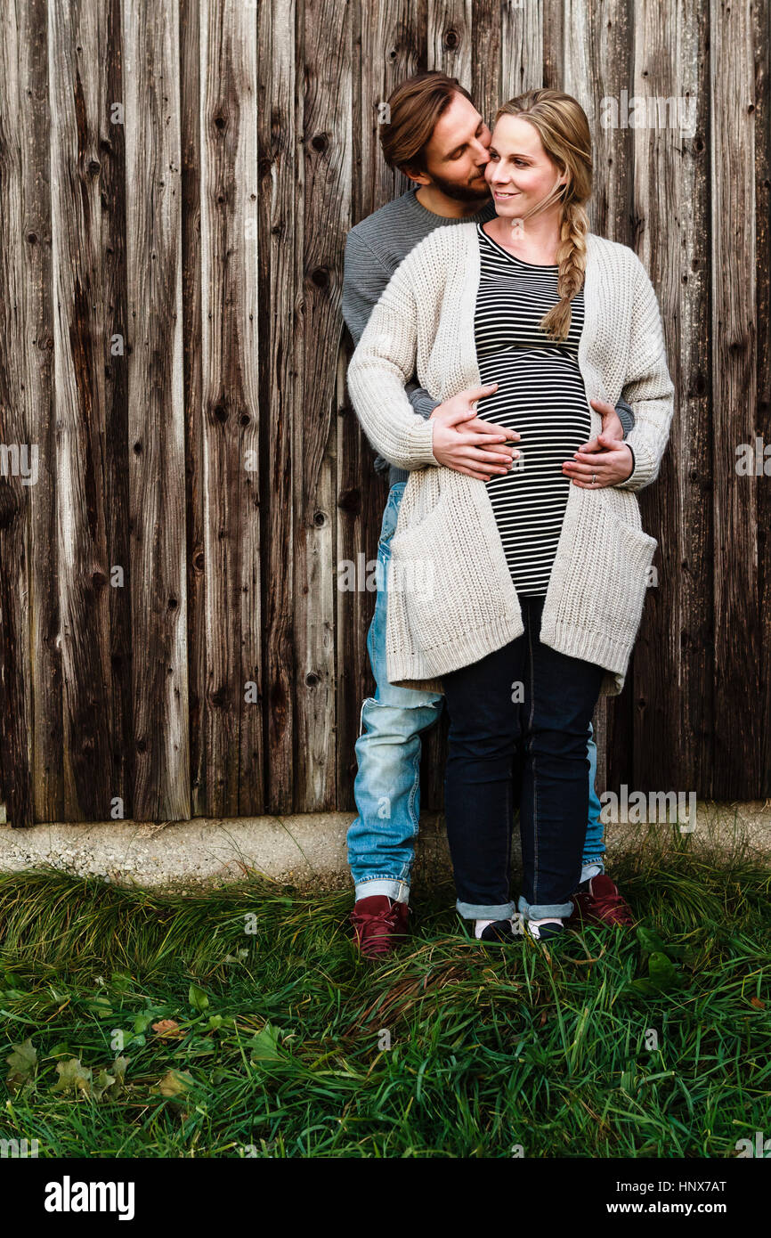 Man kissing pregnant girlfriend on cheek by wooden fence Stock Photo