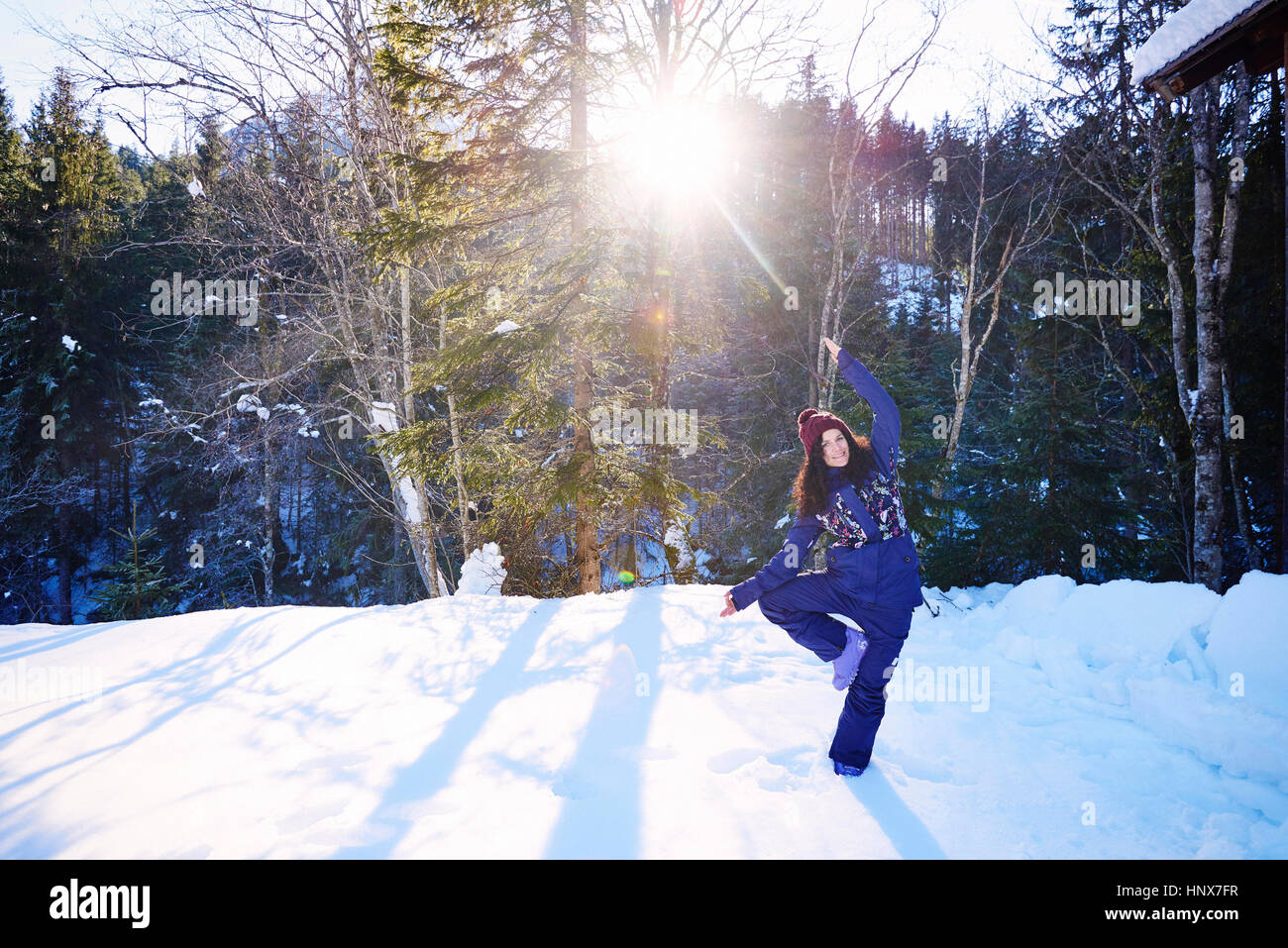 Woman in winter clothes practicing tree yoga pose in snow by forest