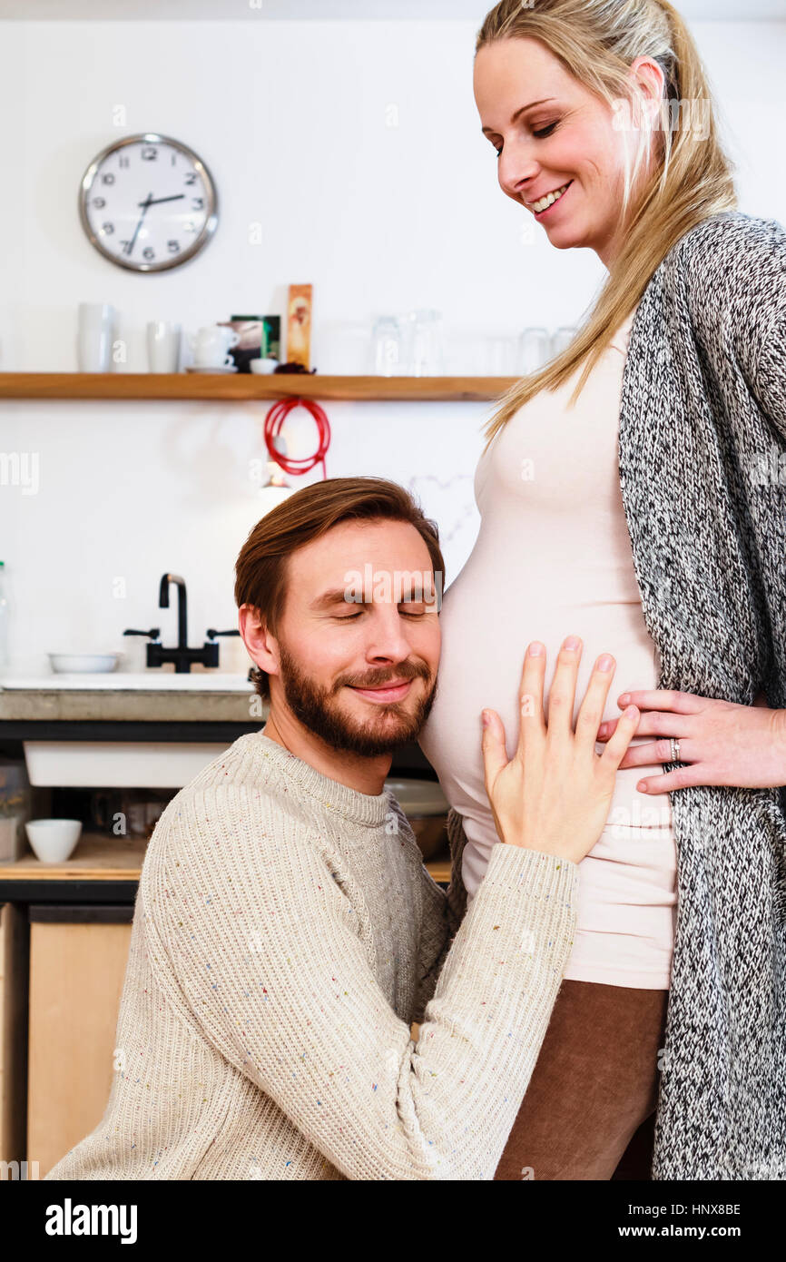 Man touching and listening to pregnant girlfriend's stomach in kitchen Stock Photo