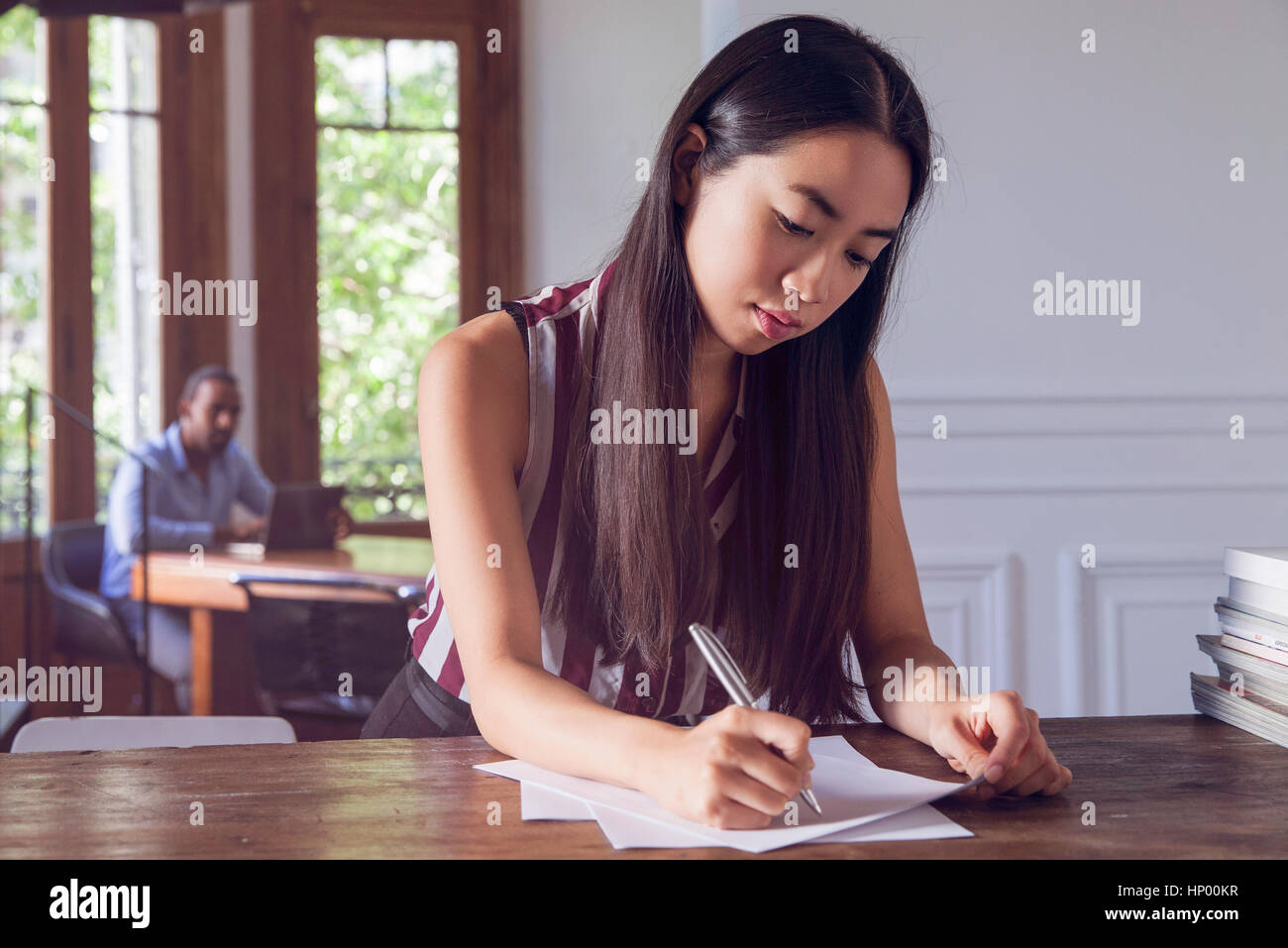 Young woman writing on paper while man watches her in the background Stock Photo