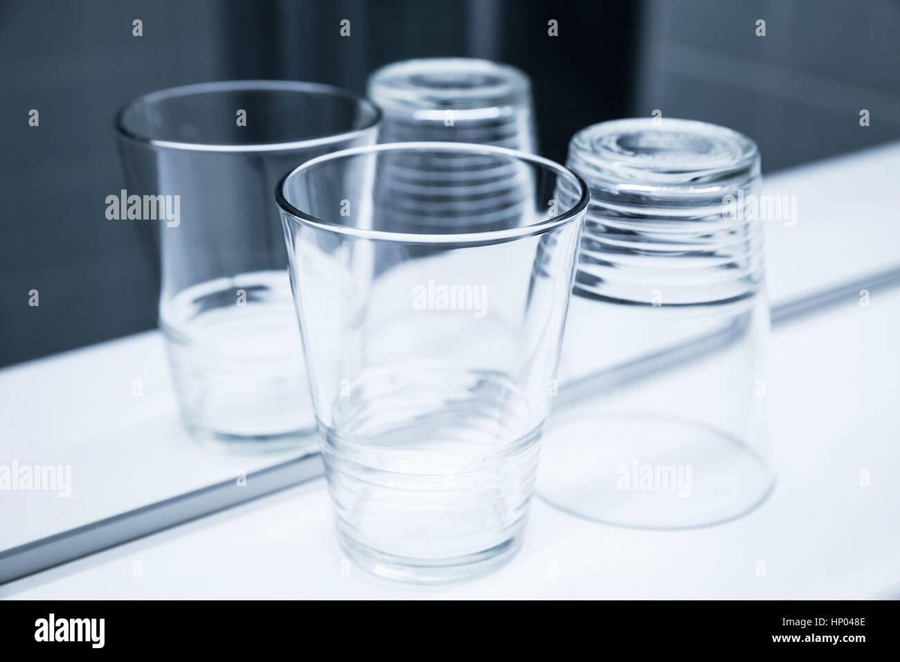 Two empty glasses stand on white shelf near the mirror, blue toned closeup photo Stock Photo
