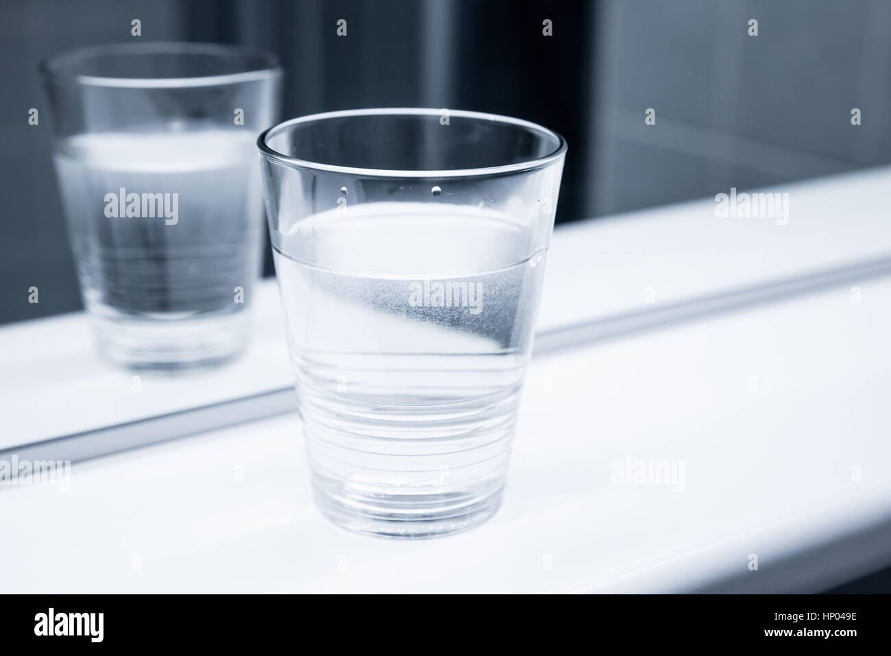 Glass of water stand on white shelf near the mirror, blue toned close-up photo Stock Photo