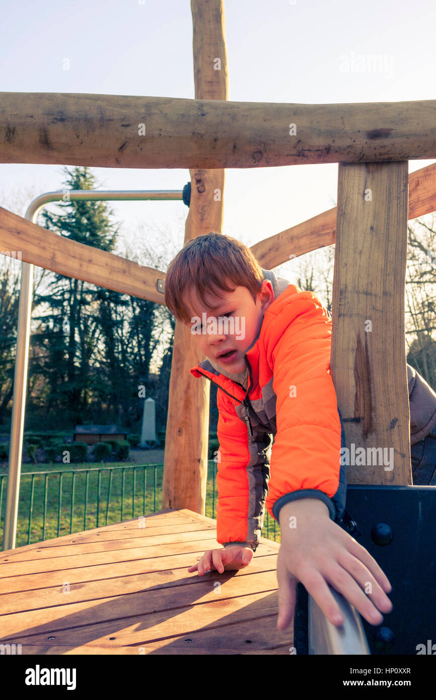 A young caucasian boy wearing a bright orange coat climbing a wooden climbing frame outdoors in a playground Stock Photo