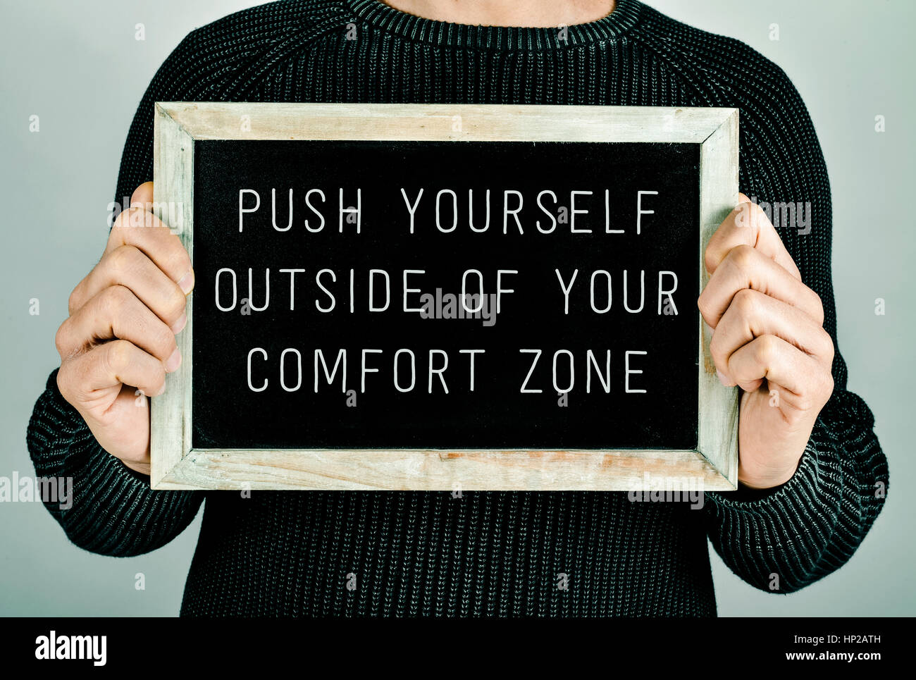 closeup of a young caucasian man holding a chalkboard in front of him with the text push yourself outside of your comfort zone written in it Stock Photo