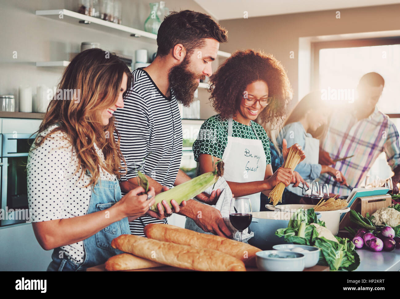Good friends laughing and talking while preparing meals at table full of vegetables and pasta ready for cooking in kitchen Stock Photo