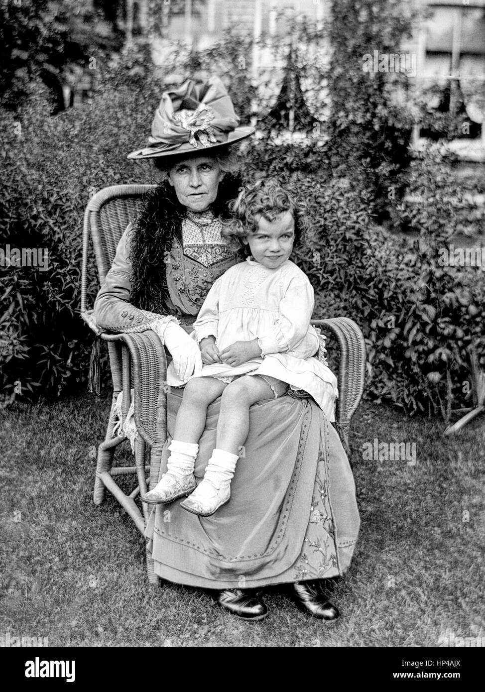 An older Edwardian lady sits in the garden in a wicker chair with a small child on her knee. The lady is wearing a long, heavy, embroidered dress, a large hat and a fur stole. The small child is wearing a short light coloured dress, short socks and sandals. Photograph taken circa 1910, restored from a high resolution scan taken from the original negative. Stock Photo