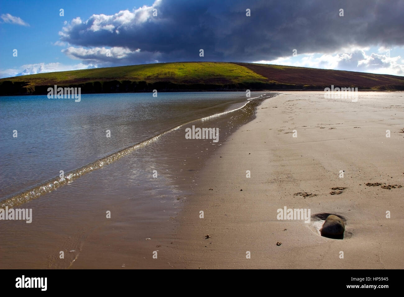 A view of the beach at Waulkmill Bay Orkney. With a dark cloud and blue sky, illuminating a green hill in the distance. With the light glinting off th Stock Photo