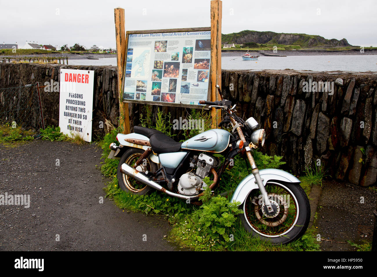 An apparently abandoned motor cycle, next to a sign for The Firth of Lorn marine special area of conservation and a danger sign. Stock Photo