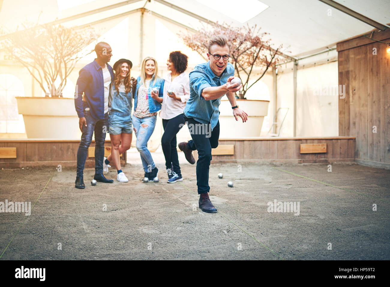 Young handsome man in glasses throwing steel petanque ball, his friends smiling standng behind