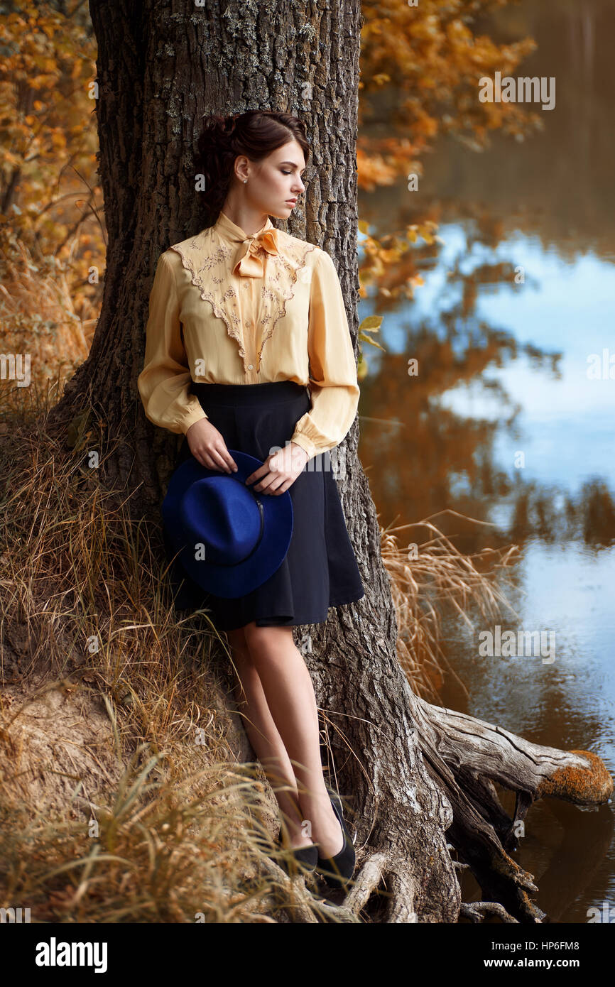 Beautiful female dreaming in the autumn park. Young brunette woman in elegant vintage blouse with bow and skirt with fashionable hat in hands. Woman w Stock Photo