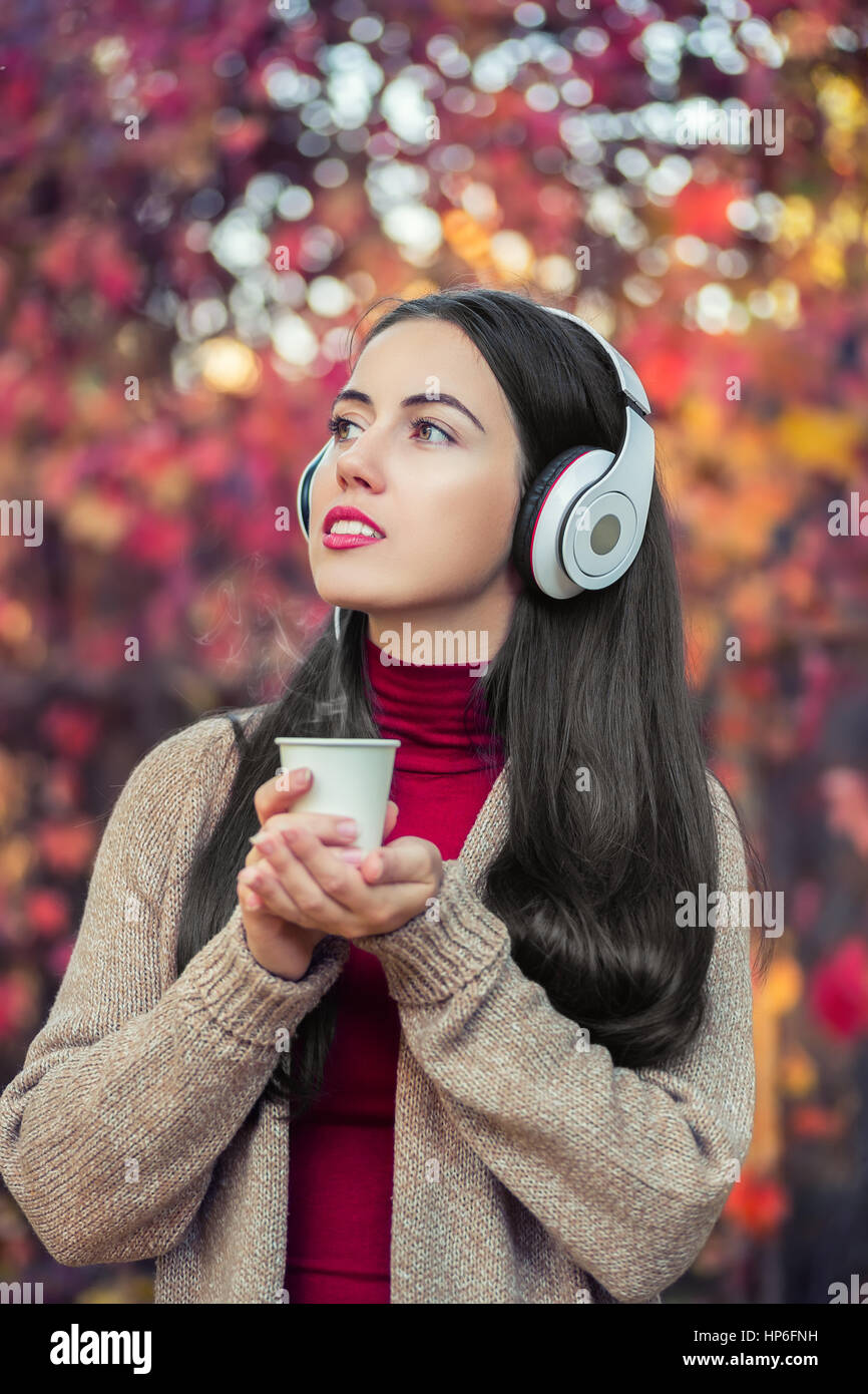 girl in autumn park with takeaway coffee and headphones. Beautiful young woman with music headphones, holding a take away coffee cup and listening to  Stock Photo
