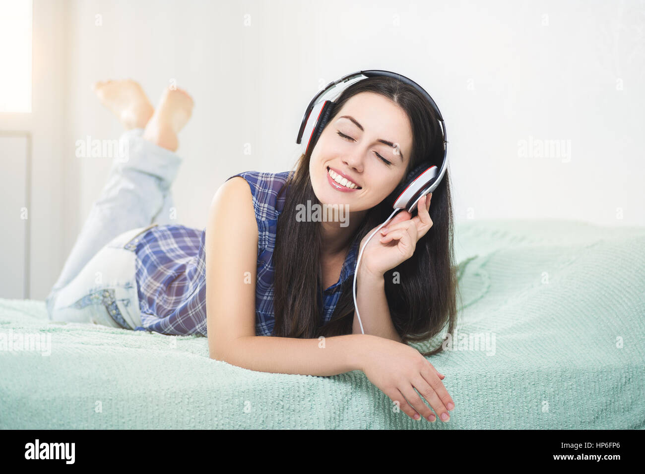 Happy relaxed girl listening music using headphones lying in bedroom with sunshine. People, leisure and technology concept - happy woman Stock Photo