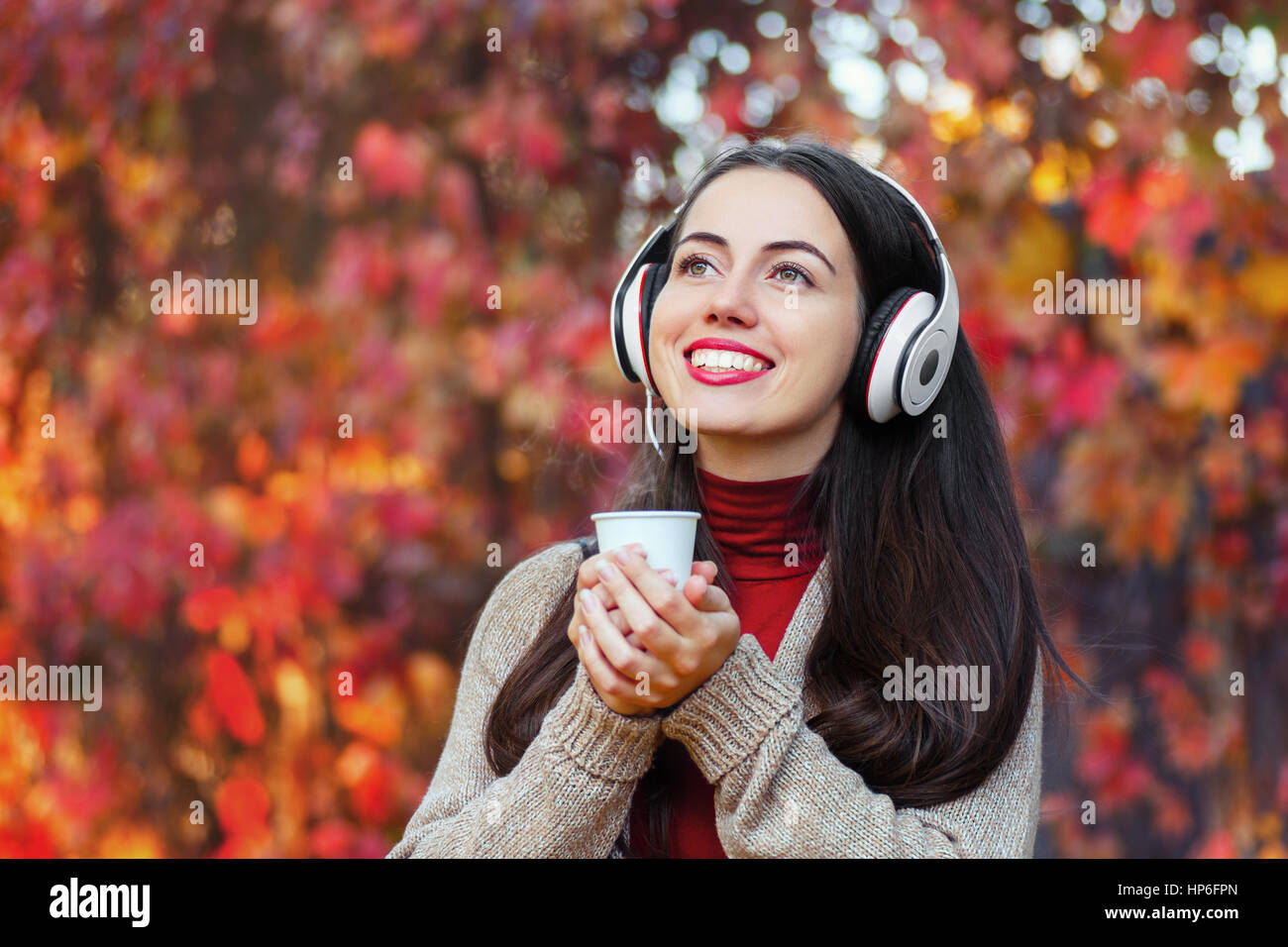 portrait of happy girl in colorful autumn park with takeaway coffee and headphones, copy space. Beautiful young woman with music headphones, holding a Stock Photo
