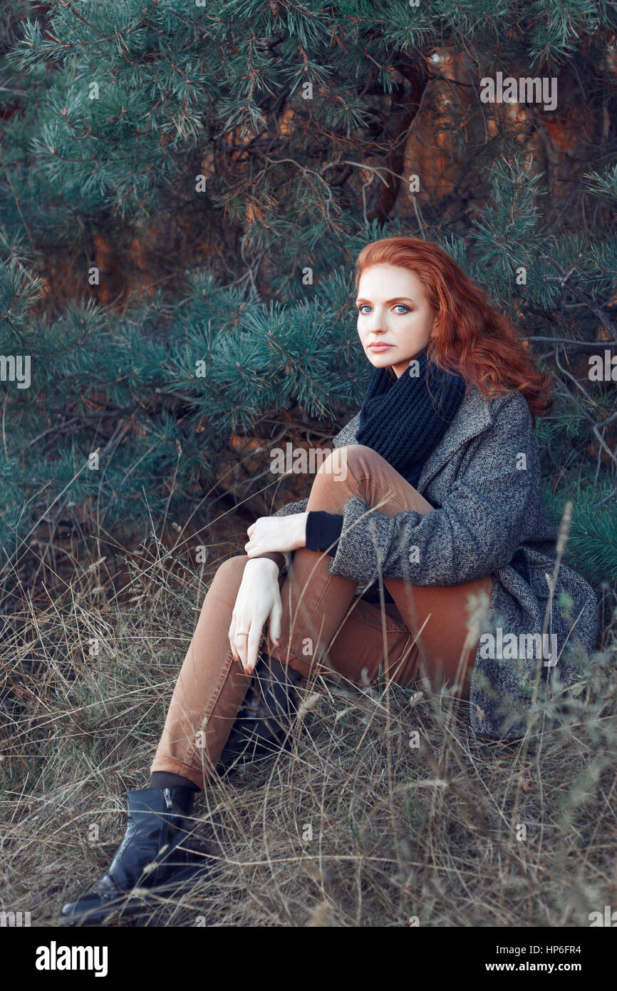 Beautiful elegant red-haired woman sitting in park in autumn. Redhead girl in autumn clothes in the forest. Redhead woman outdoor in autumn park, wear Stock Photo