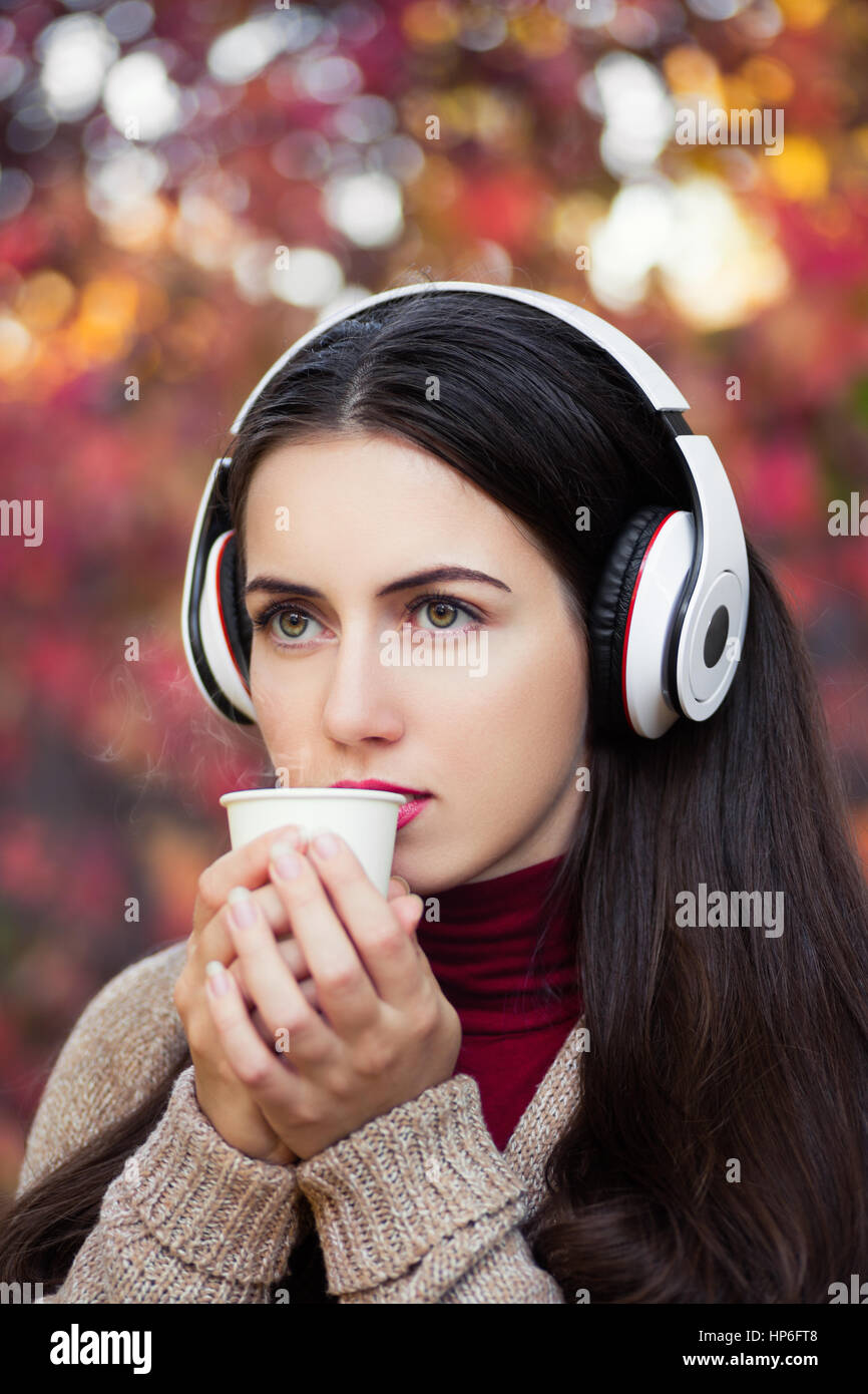 autumn woman drinking hot coffee in fall city park or forest beautiful fall colors. Close-up portrait of beautiful young woman with music headphones Stock Photo