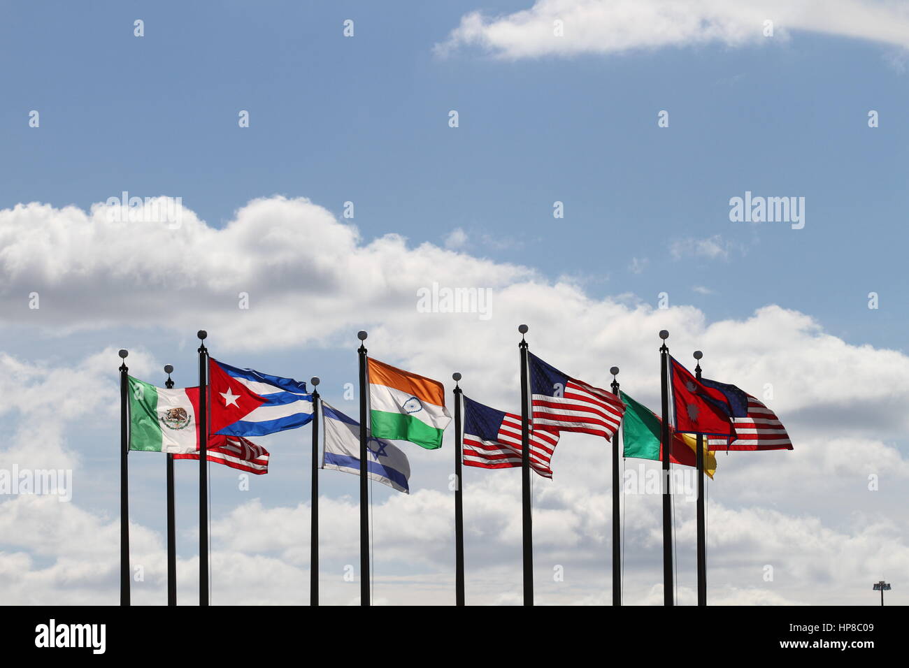 The flags US, Nepal, Cameroon,  Israel, India, Mexico and Cuba on a flagpoles blowing in the wind against a blue sky and white clouds. The wind blows. Stock Photo