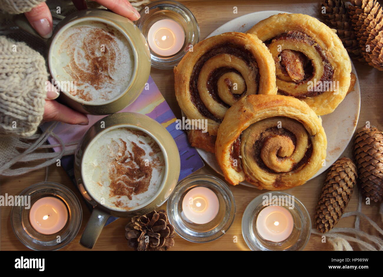 A woman reaches for a creamy mocha (coffee and chocolate) served with cinnamon buns) served by candle light in a cosy English home in winter Stock Photo