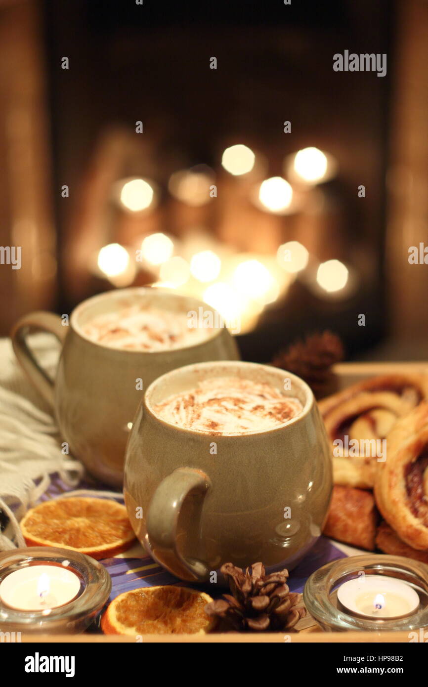 Cosy concept. Hygge. Creamy mugs of mocha - coffee and chocolate - are served with cinnamon buns by a candle lit open fireplace in a cosy English home Stock Photo