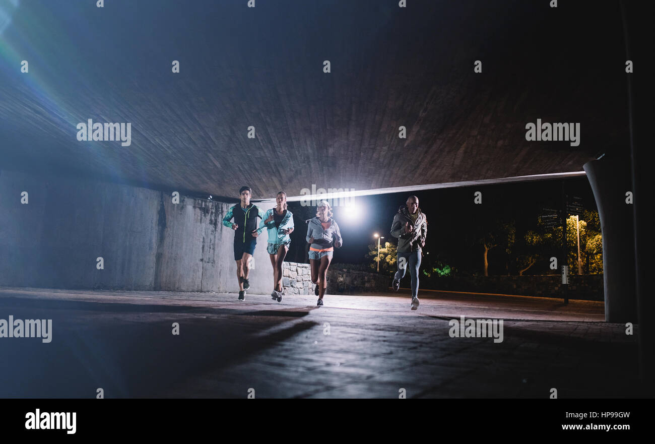 Group of runners running under a bridge in city. Young men and women jogging together at night. Stock Photo