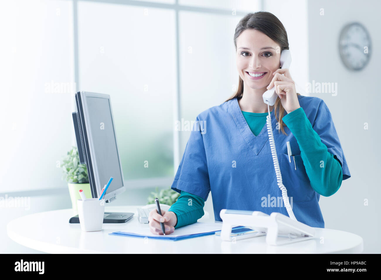 Young female doctor and practitioner working at the reception desk, she is answering phone calls and scheduling appointments Stock Photo