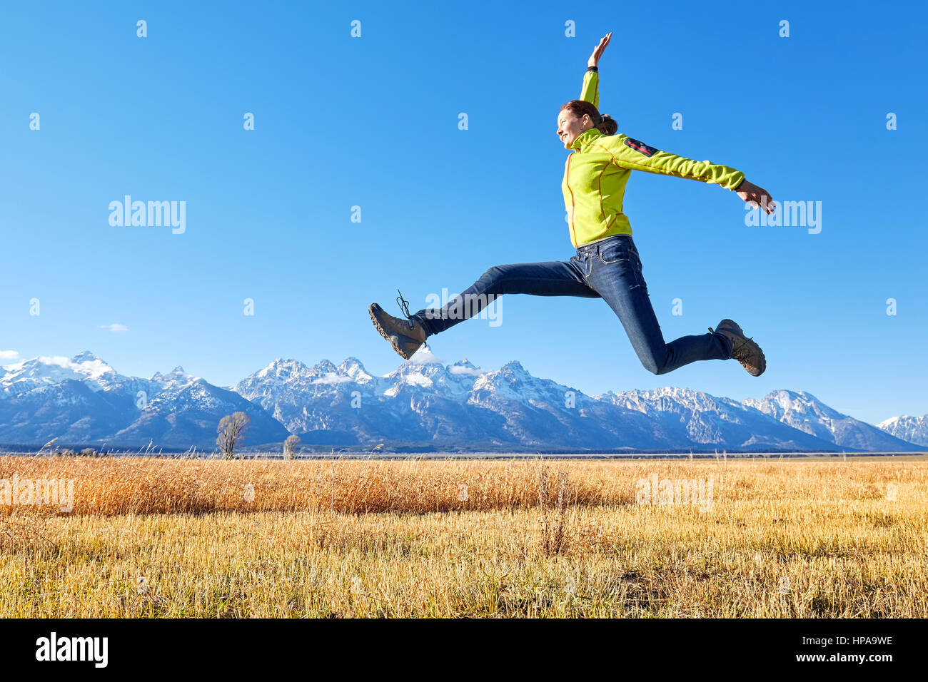 Happy young woman jumping on a meadow, Grand Teton mountain range in distance, Wyoming, USA. Stock Photo
