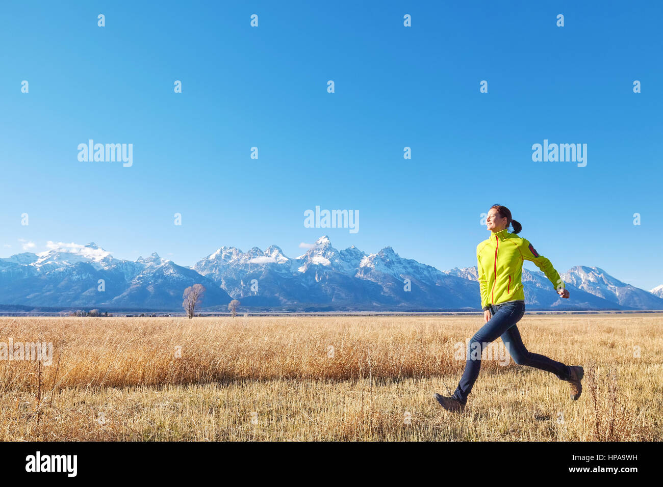 Happy young woman runs on a meadow at sunset, Grand Teton mountain range in distance, Grand Teton National Park, Wyoming, USA. Stock Photo