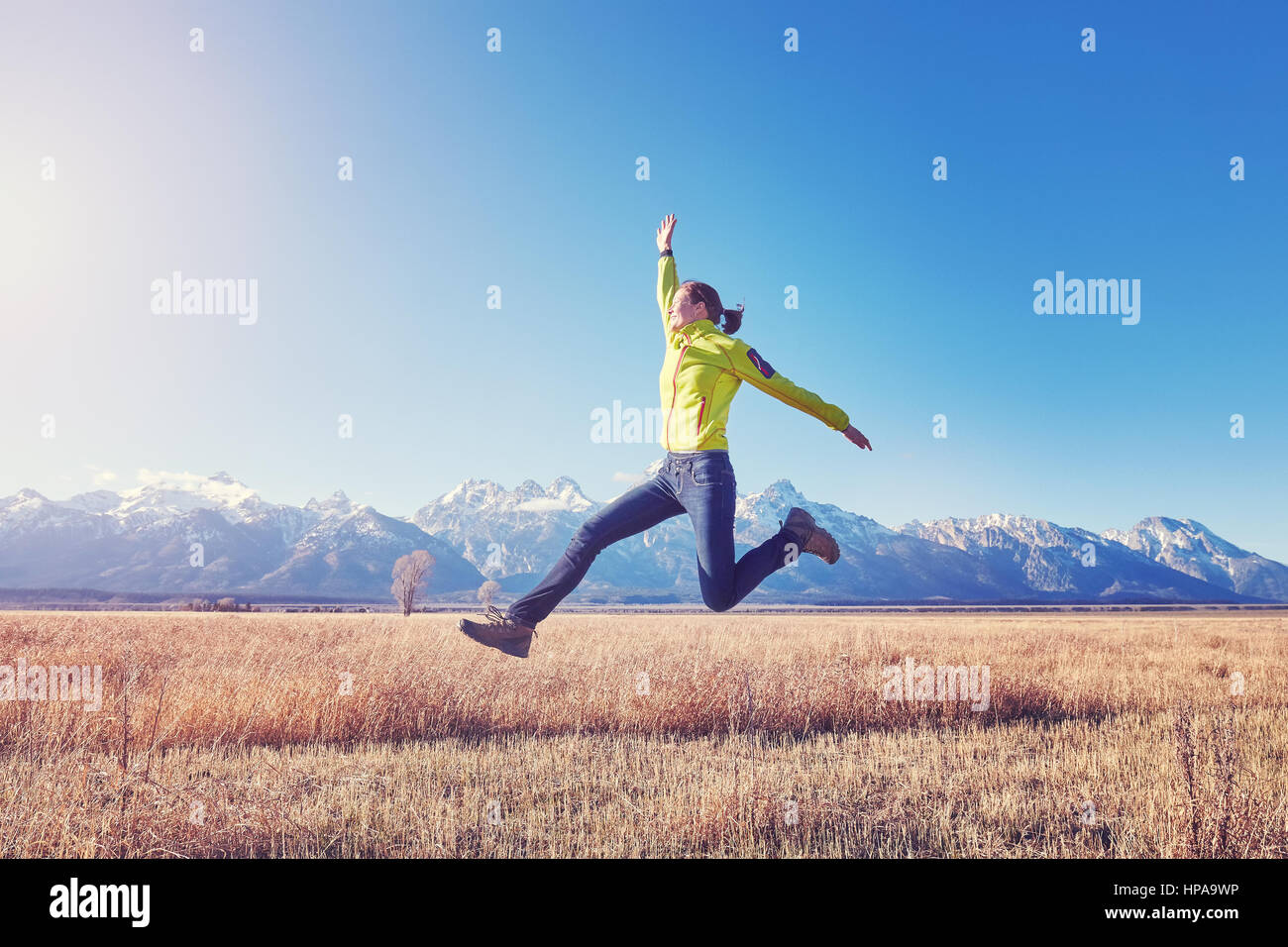 Happy young woman jumping on a meadow at sunset, color toned image, Grand Teton mountain range in distance, Wyoming, USA. Stock Photo
